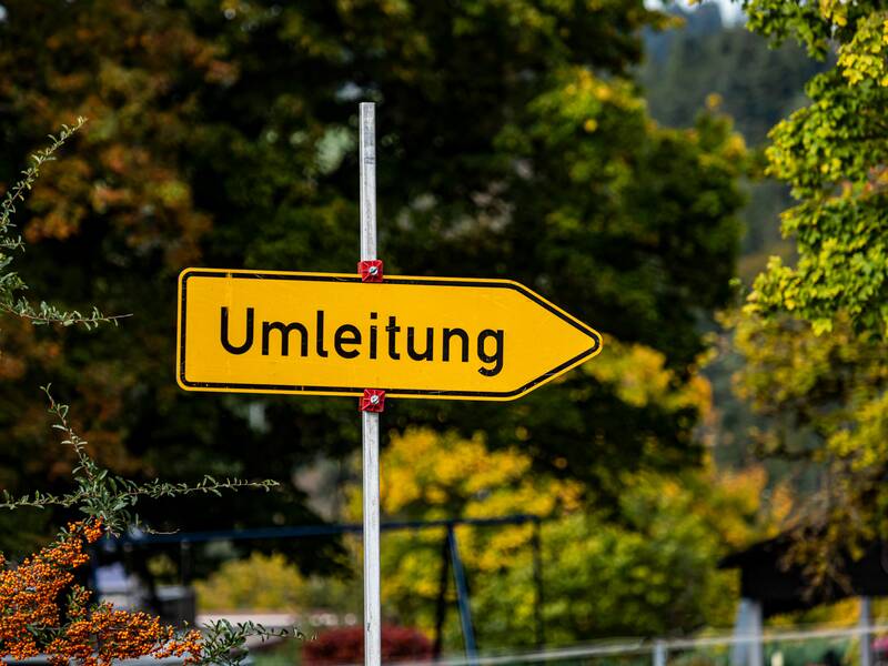 Ein gelbes Umleitungsschild, das in die rechte Richtung zeigt. Im Hintergrund sind herbstliche Bäume uns Sträucher.